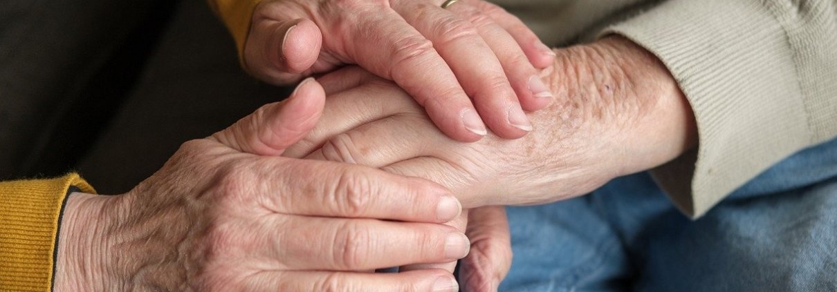 Ältere Frau hält Hand ihres pflegebedürftigen Manns. Nur die Hände sind zusehen.