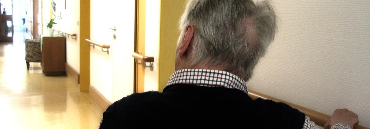 Ältere Mann im Flur eines Pflegeheims und sitzt im Rollstuhl.