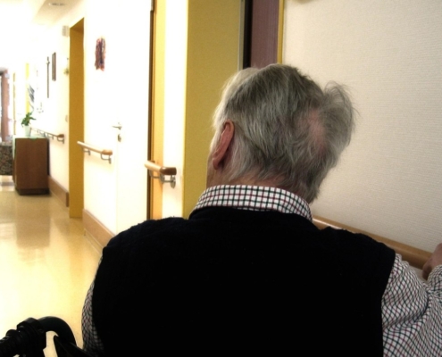 Ältere Mann im Flur eines Pflegeheims und sitzt im Rollstuhl.