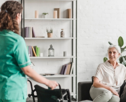 Pflegekraft, die Rollstuhl zu ihrer Patientin bringt, Patientin sitzt auf der Couch.
