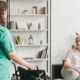 Pflegekraft, die Rollstuhl zu ihrer Patientin bringt, Patientin sitzt auf der Couch.
